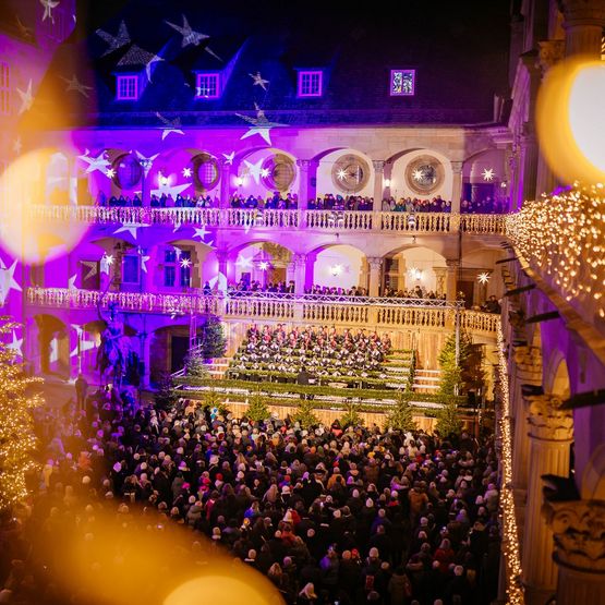 Blick auf feierlich erleuchteten Innenhof des Alten Schlosses mit Weihnachtsbaum und Chorknaben auf der Bühne. Schöne Lichtreflexionen und weihnachtliche Stimmung.